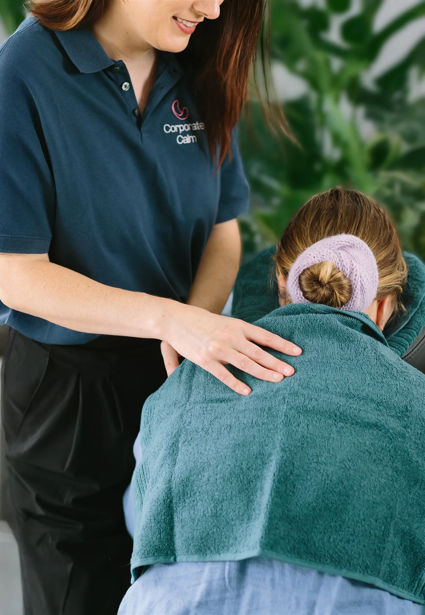 A therapist in a Corporate Calm shirt giving a shoulder massage to a client covered with a green towel and wearing a lavender hair scrunchie.