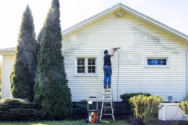 Man standing on a ladder pressure washing the exterior wall of a white house.