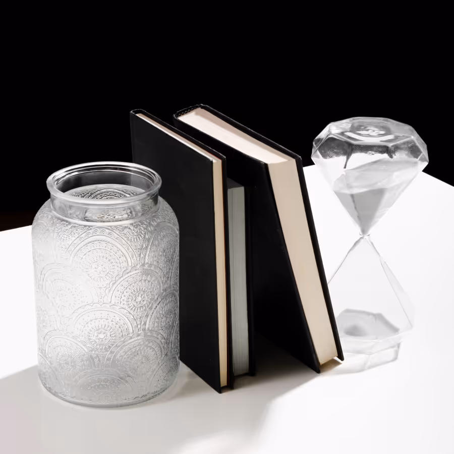 Two black-covered books standing upright between a patterned glass jar and a diamond-shaped hourglass on a white surface with a black background.