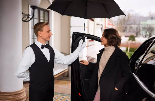 A valet in formal attire holding an umbrella over a woman stepping out of a car outside a building.