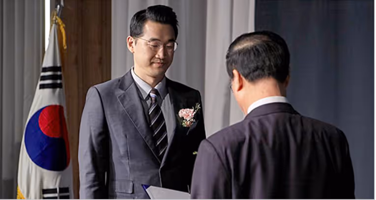 Man with glasses receiving an award or certificate from another man in a formal setting with a South Korean flag in the background.