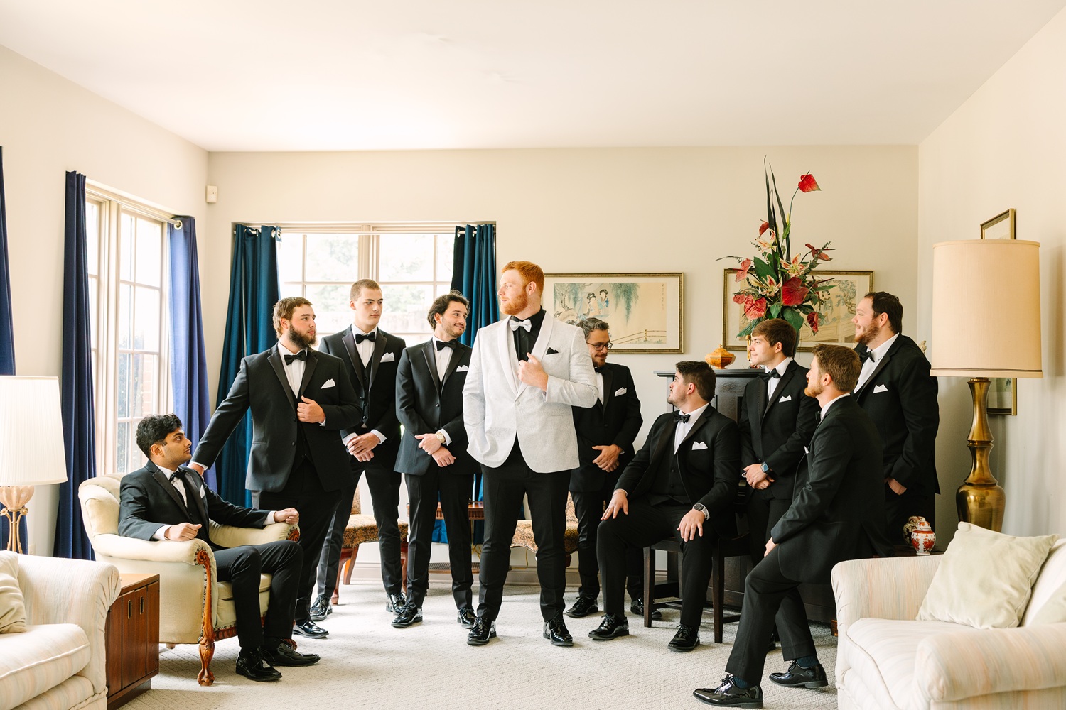 A group of ten men dressed in black tuxedos with bow ties; one man in a white tuxedo jacket stands in the center, posing in a well-lit living room.