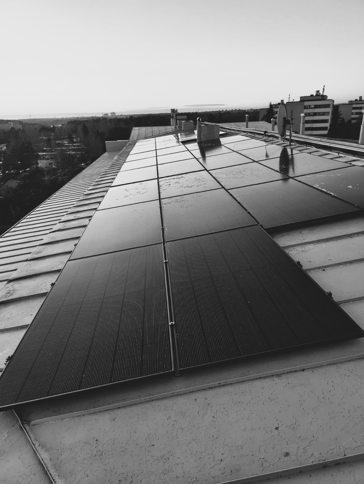 Ralos-aurinkopaneelit asennettuna kaltevalle katolle, taustalla kaupunkimaisema ja puita kirkkaan taivaan alla.