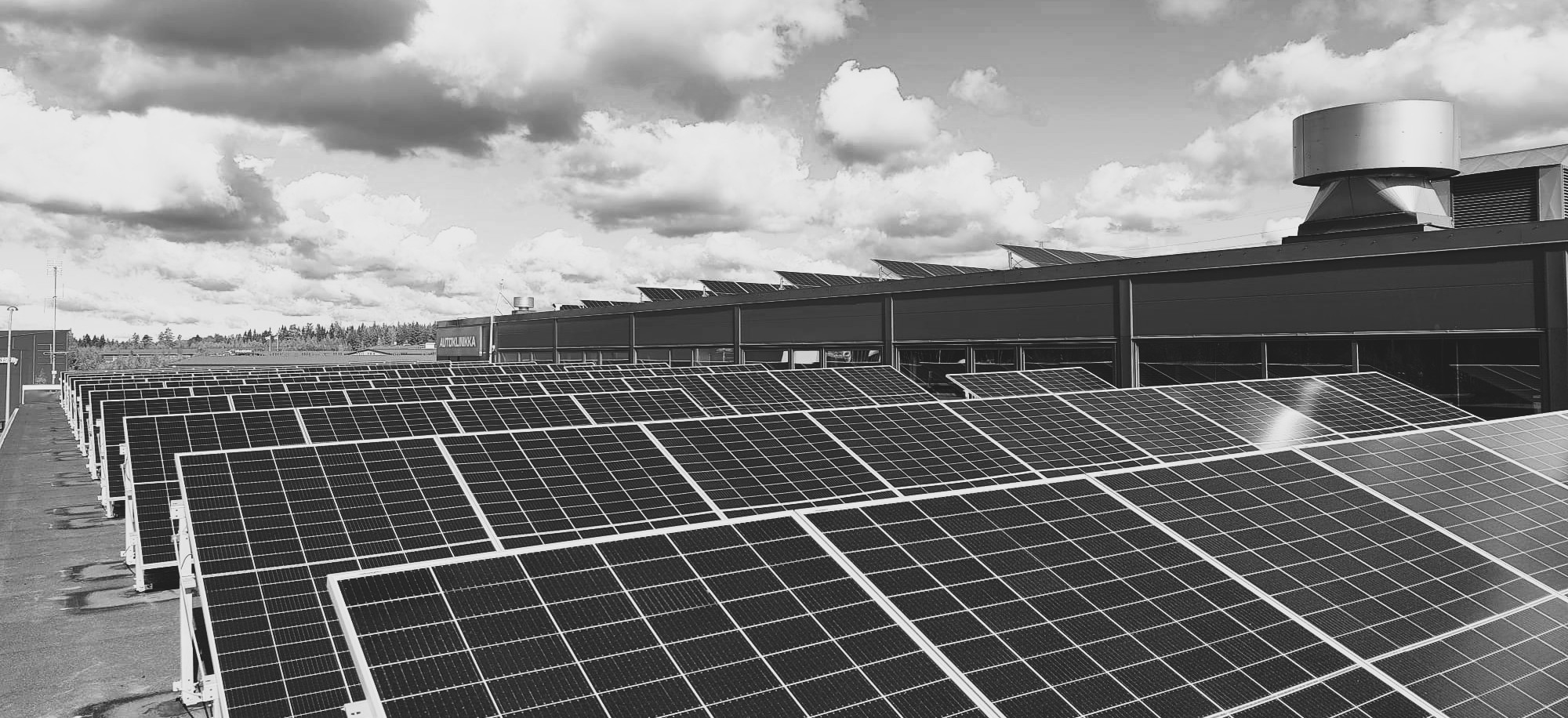 Riveittäin asennettuja Ralos-aurinkopaneeleja tasakatolla osittain pilvisen taivaan alla.