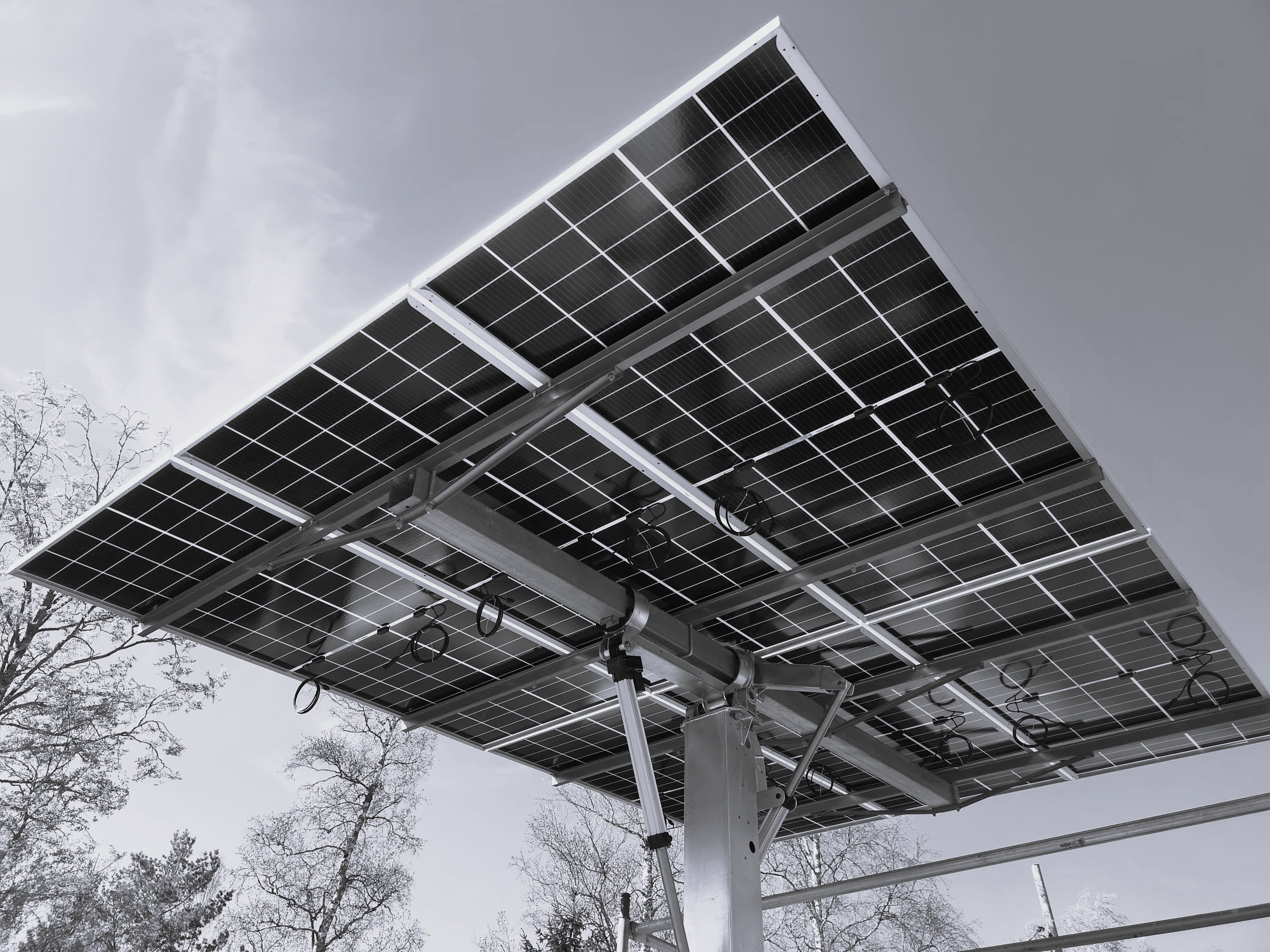 Näkymä Ralos-aurinkopaneelirivistön alapuolelta metallisella tukirakenteella, taustalla puita ja kirkas taivas.