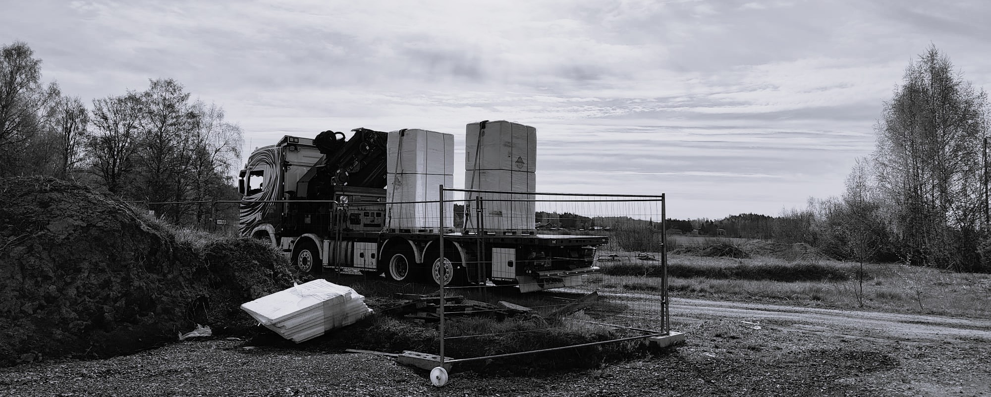 Kuorma-auto kuljettamassa paketoituja kuormalavoja työmaalla, taustalla puita ja pilvinen taivas.