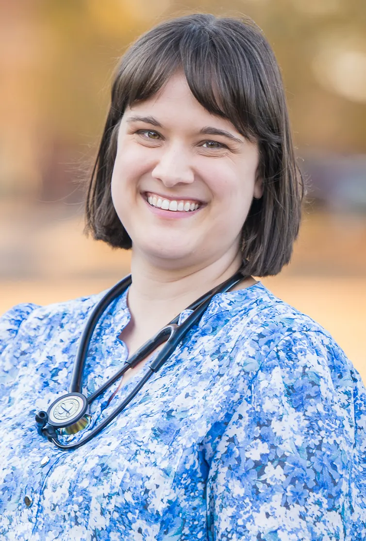 Smiling woman wearing a blue floral blouse with a stethoscope around her neck against a blurred outdoor background.