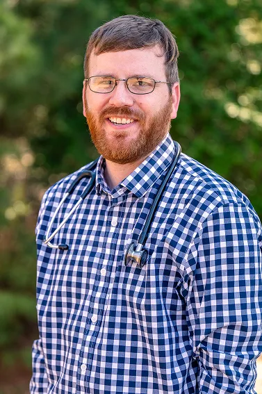 Smiling male doctor with glasses and beard wearing a blue checkered shirt and stethoscope around his neck in an outdoor setting.