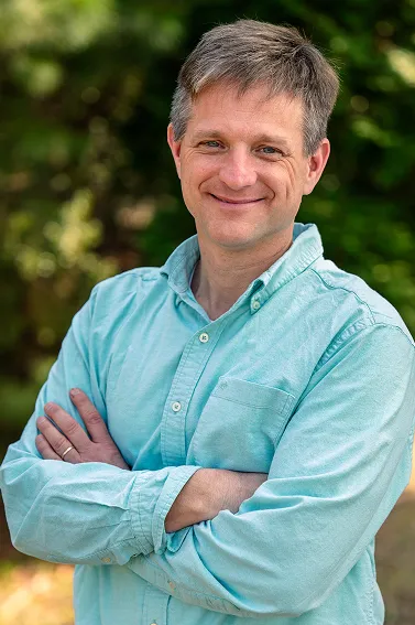 Smiling man with grayish hair wearing a light blue button-up shirt, standing outdoors with arms crossed.