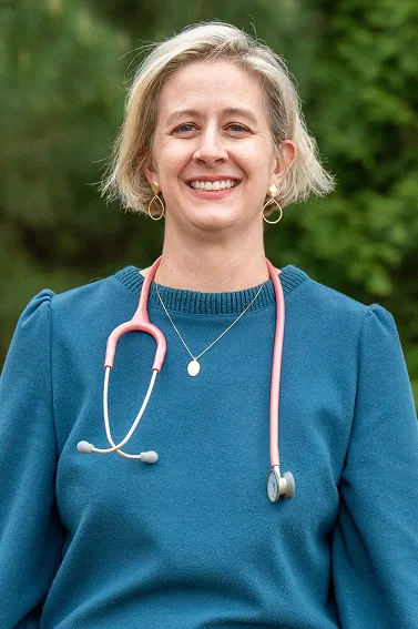 Smiling woman with short blonde hair wearing a teal sweater and a pink stethoscope around her neck, standing outdoors with a green blurred background.