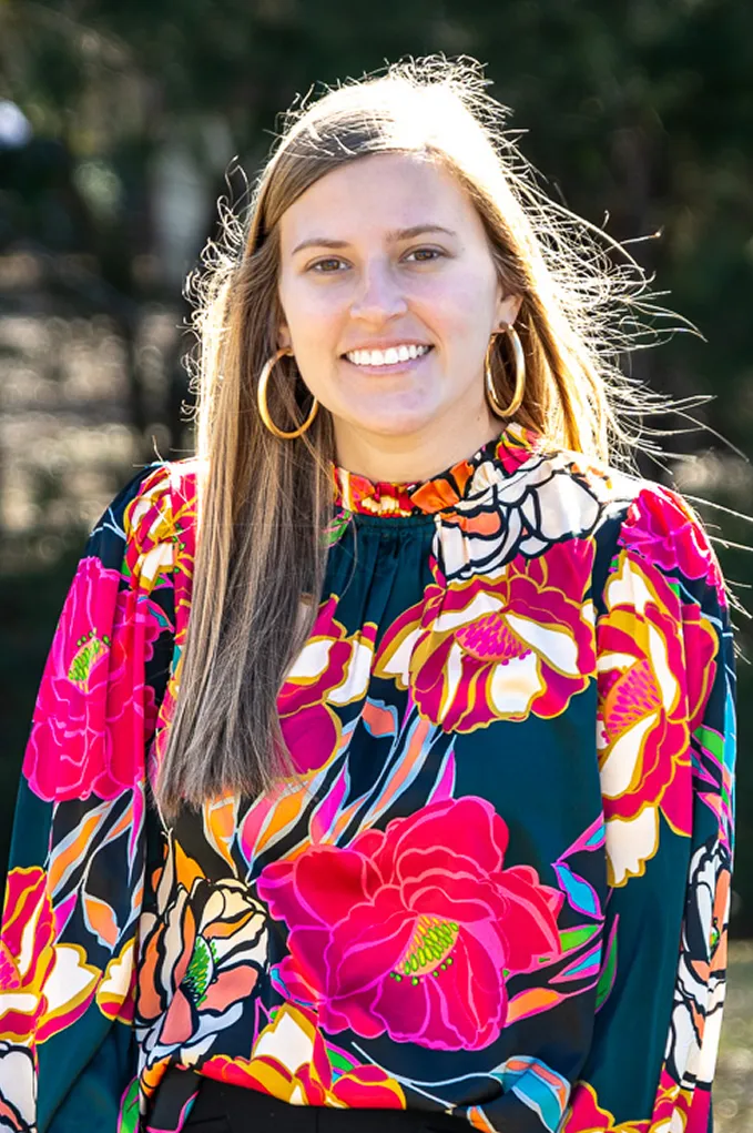 Smiling woman with long light brown hair wearing large hoop earrings and a colorful floral blouse outdoors.