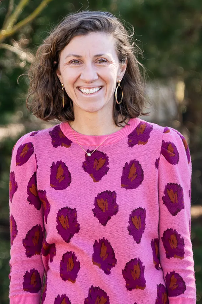 Smiling woman with shoulder-length brown hair wearing a pink sweater with purple and brown leaf patterns, standing outdoors.