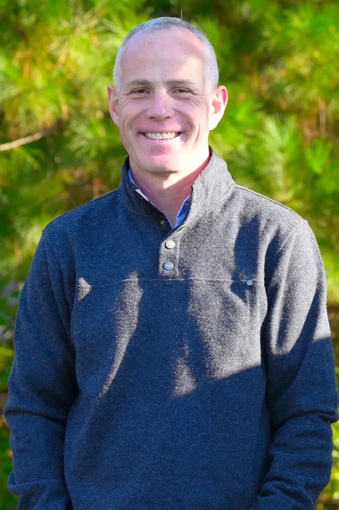 Smiling man with short gray hair wearing a dark gray pullover standing outdoors with green foliage background.