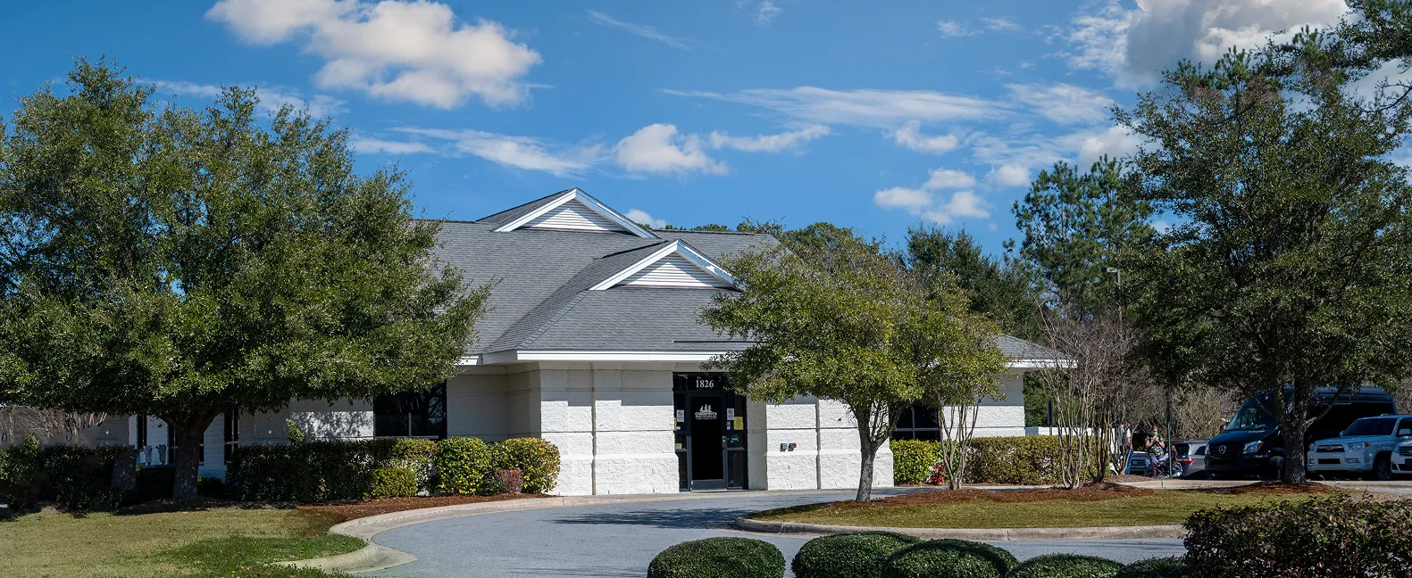 A single-story white office building with a gray roof surrounded by trees and shrubs under a blue sky with clouds.