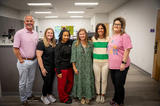 Group of six smiling people standing in a modern indoor space with light walls and flooring.