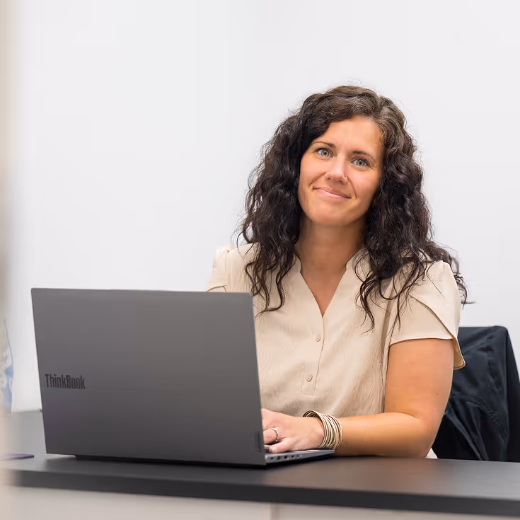 Woman with curly dark hair smiling while sitting at a desk and using a Lenovo ThinkBook laptop.