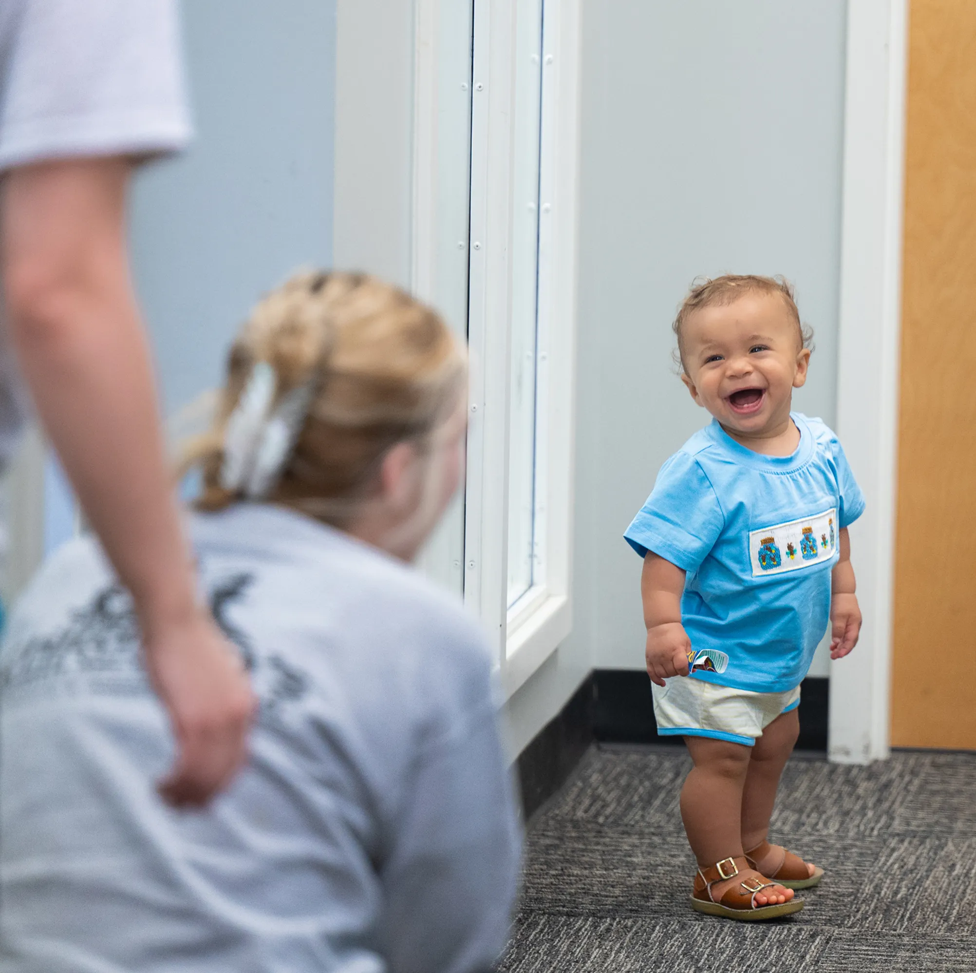 Smiling toddler in a blue shirt and sandals standing indoors near a door while a woman crouches nearby.