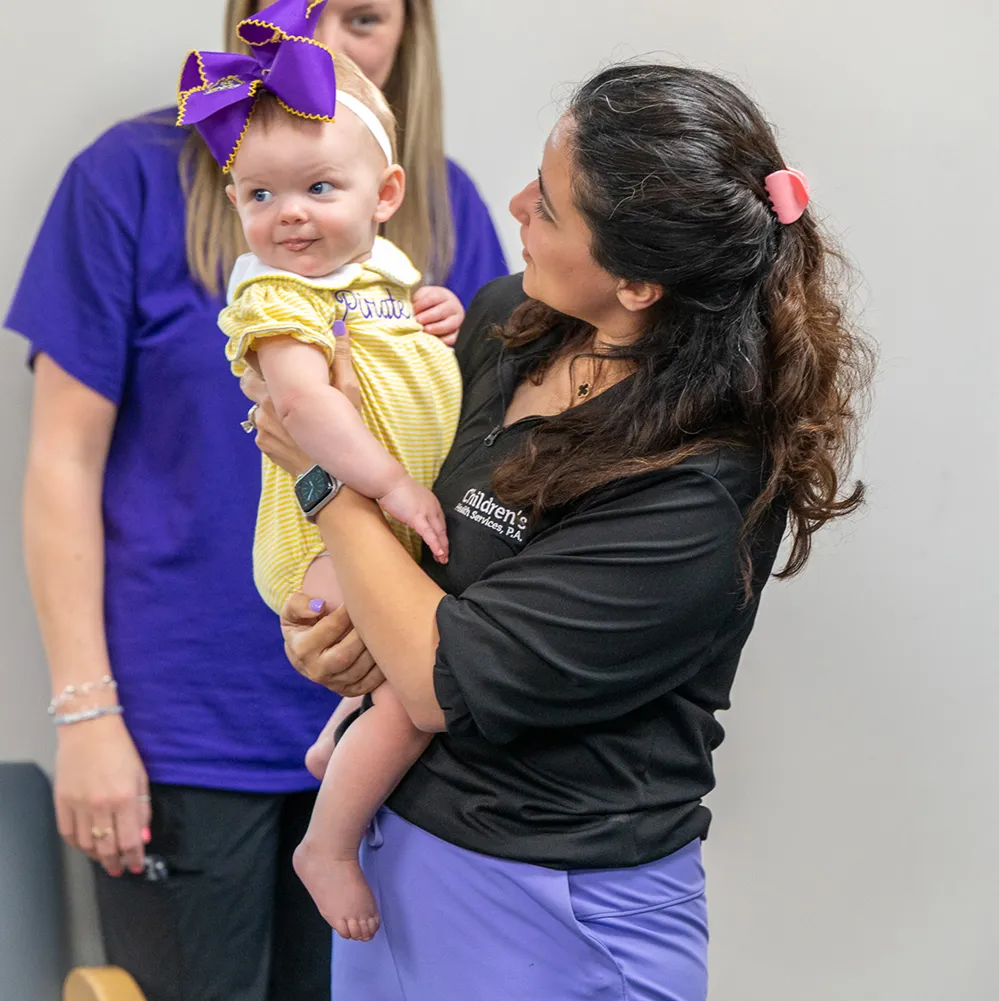 Woman in a black shirt holding a baby wearing a yellow striped outfit and a large purple bow, with another person in purple in the background.