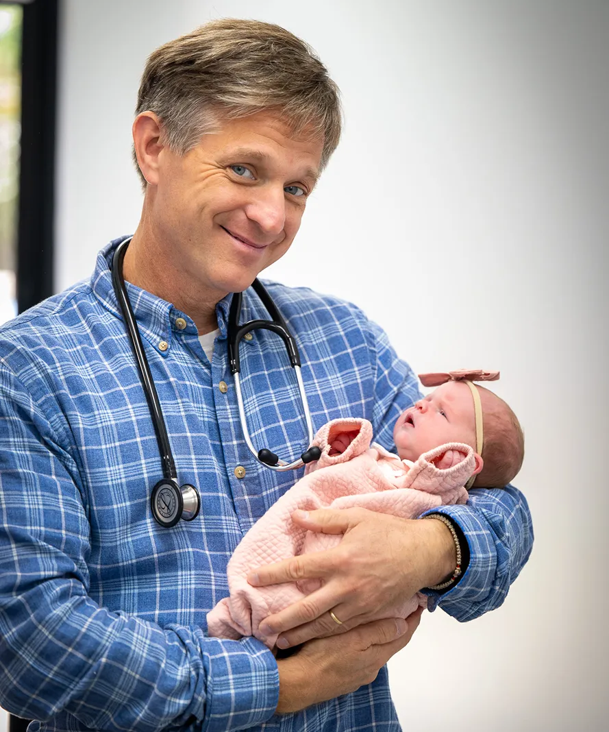Male doctor in blue plaid shirt holding a newborn baby dressed in pink with a headband.