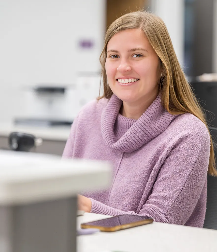 Smiling young woman wearing a lavender sweater sitting at a table with a smartphone in front of her.