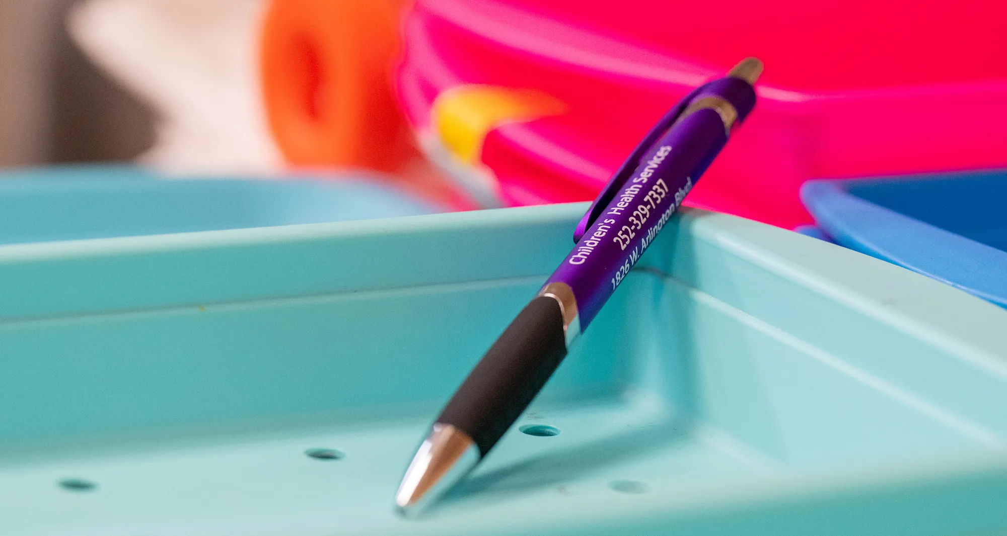 Purple pen with text 'Children's Health Services' resting diagonally inside a light blue plastic tray with blurred colorful trays in the background.