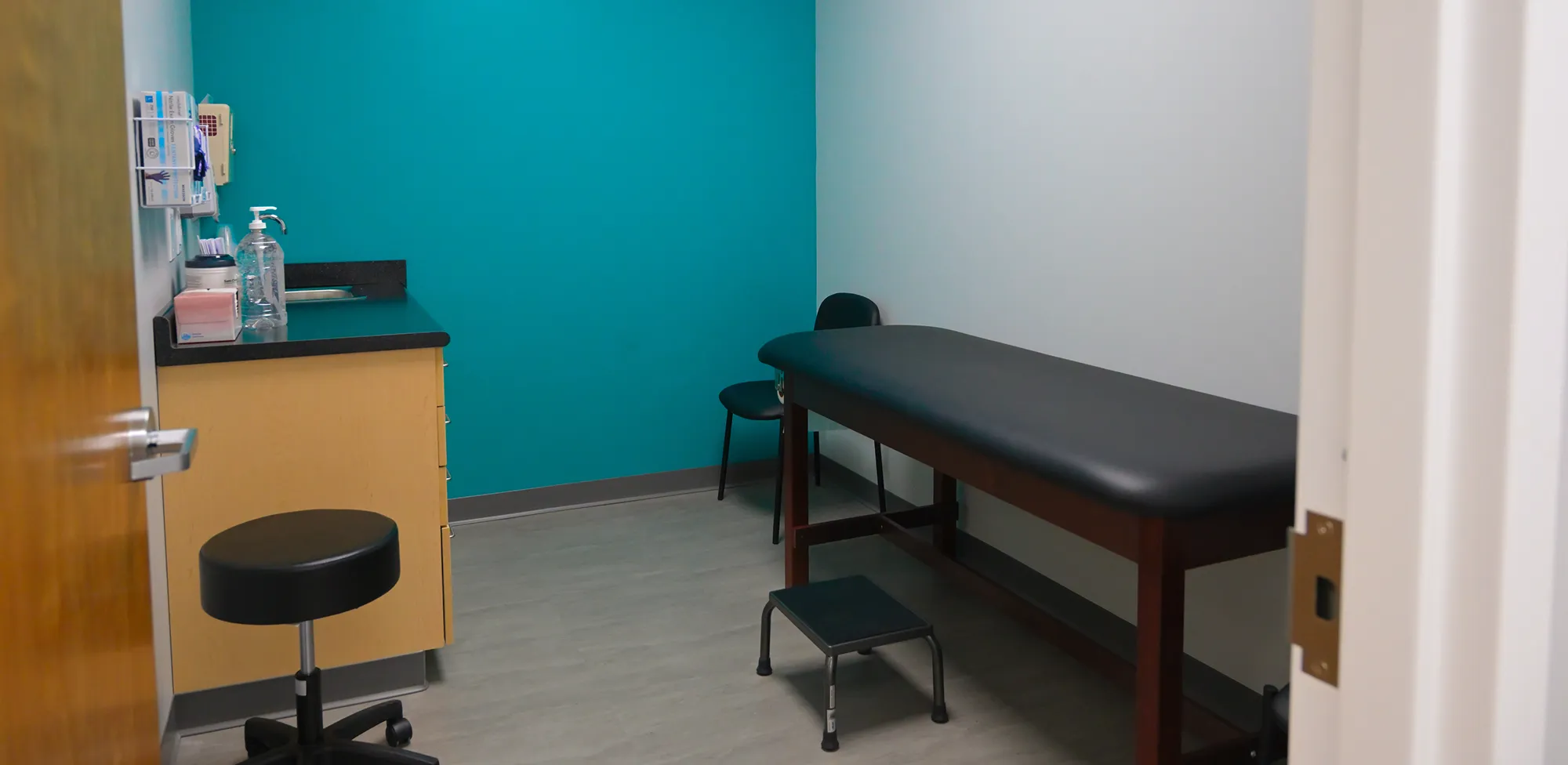 Empty medical examination room with a black exam table, small step stool, two chairs, wooden cabinet, and bright teal accent wall.