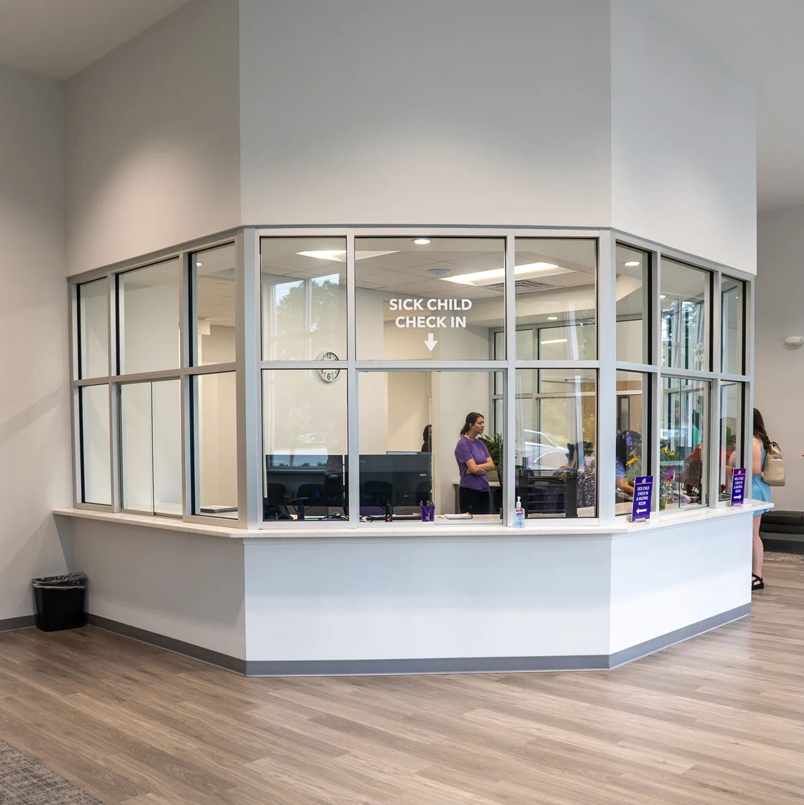 Reception area with glass windows labeled 'Sick Child Check In,' showing staff and visitors inside a medical facility.