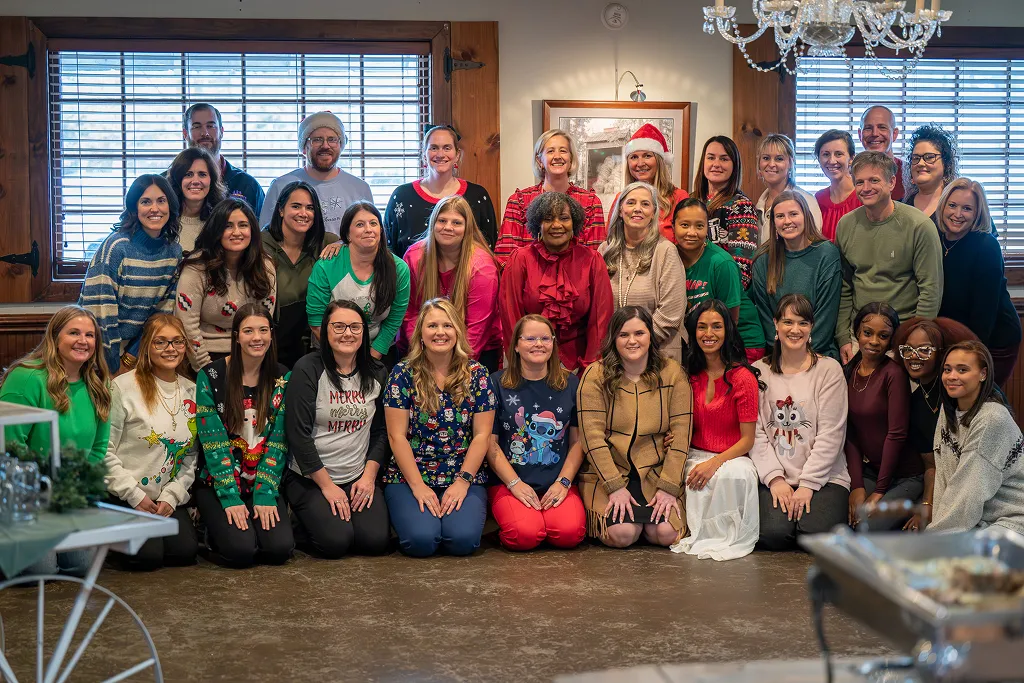 Group of 31 people posing indoors in front of windows wearing festive holiday sweaters and clothing.