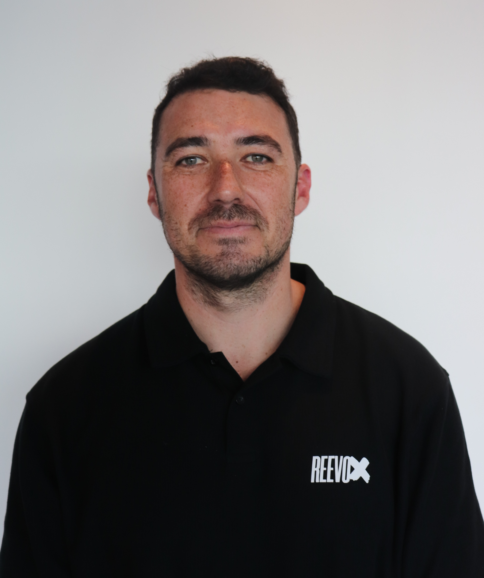 Man with short dark hair and stubble wearing a black polo shirt with a REEVOX logo on a white background.