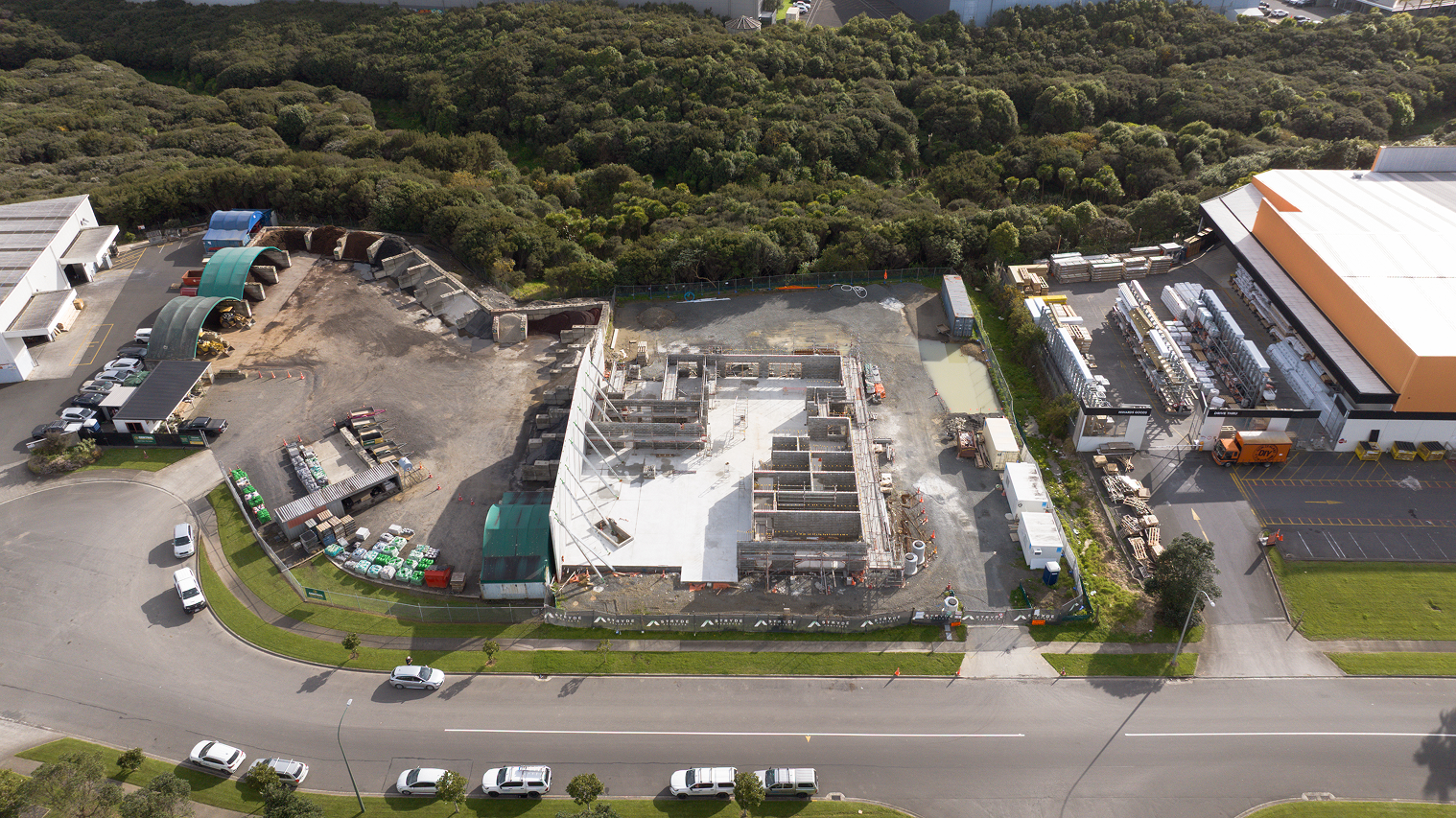 Aerial view of a construction site with partially built concrete structures surrounded by a fence, adjacent to a warehouse and dense green forest in the background.
