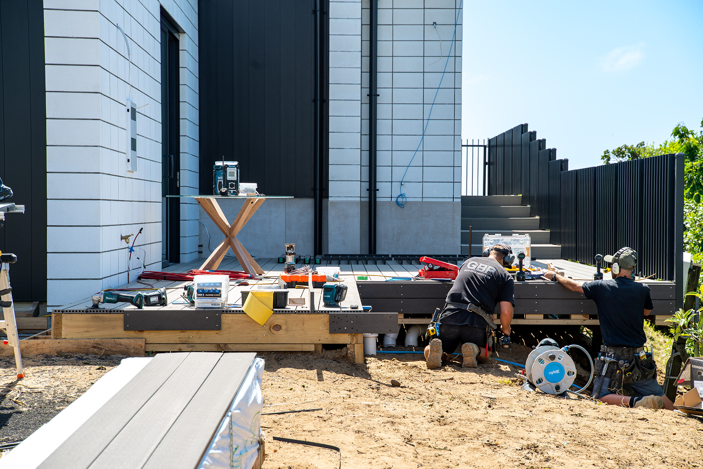 Two construction workers installing decking on a wooden patio outside a modern building with white tiled walls and black fencing.