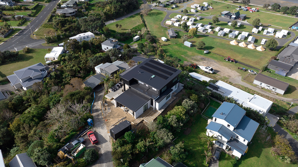 Aerial view of a modern black and white house under construction surrounded by greenery, nearby homes, and a campsite with tents and caravans.