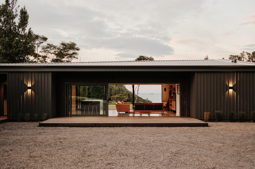 Modern house with dark metal siding, a wooden deck, and large open sliding doors revealing a seating area and a view of trees and water beyond.
