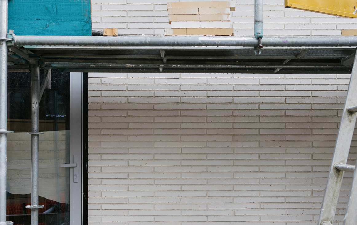Construction scaffolding and ladder in front of a beige brick wall with a partially visible door on the left.