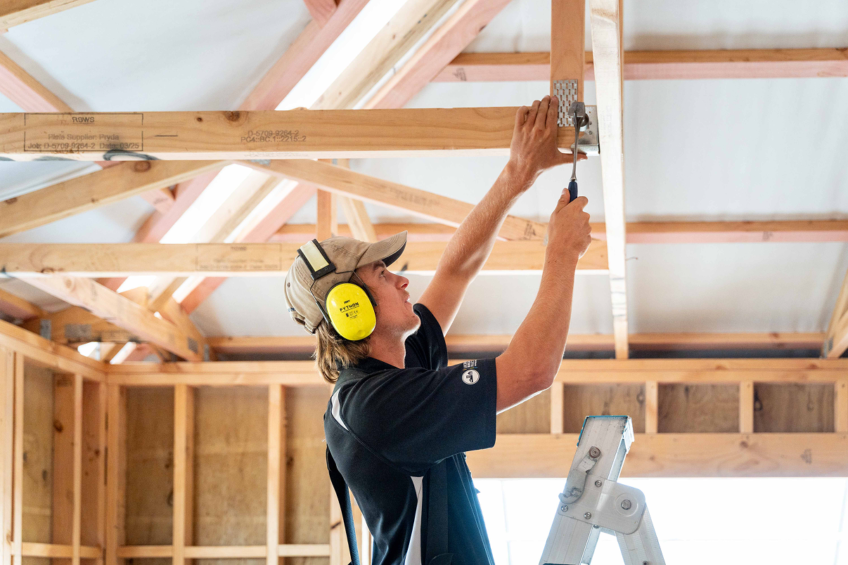 Construction worker wearing hearing protection and cap fixing a metal bracket on wooden beams indoors.