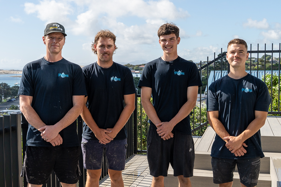 Four men wearing black extreme team t-shirts and shorts standing outdoors on a sunny day with a scenic water and hillside background.