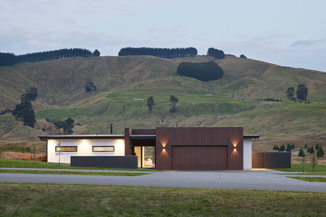Modern single-story house with white and wooden exterior set against rolling green hills at dusk.