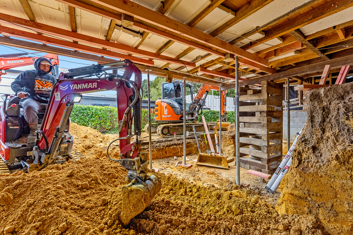 Construction worker operating a red mini excavator digging dirt under a wooden house foundation with another orange excavator and tools in the background.