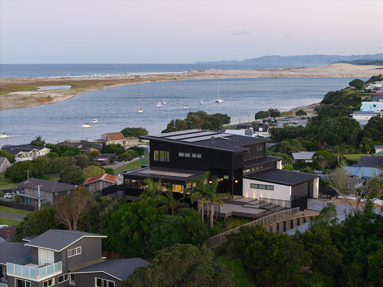 Aerial view of a modern black and white house under construction surrounded by greenery, nearby homes, and a campsite with tents and caravans.