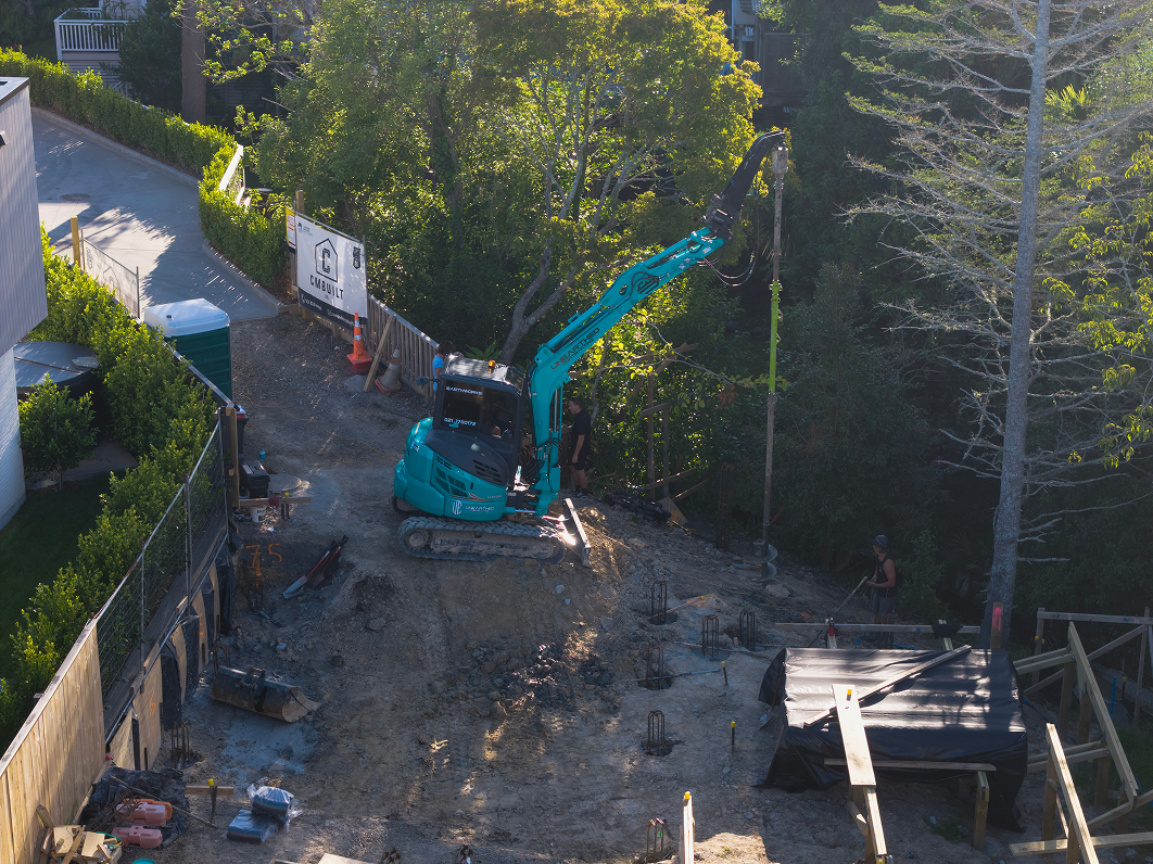 Construction worker operating a red mini excavator digging dirt under a wooden house foundation with another orange excavator and tools in the background.