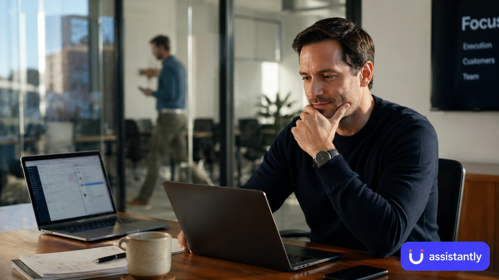This image shows a professional man sitting at a desk in a modern office environment, working intently on a laptop. The man is thoughtfully focused, with one hand resting on his chin. On the desk in front of him are an open notebook, a pen, a coffee mug, and a smartphone. Another laptop displaying a calendar or scheduling application is also on the desk. In the background, a blurred colleague is walking past a glass partition. The office features a large display screen with the word 'Focus' visible, indicating a meeting or presentation setting. This image is useful for illustrating business productivity, office work, remote team collaboration, or technology use in a professional workspace.