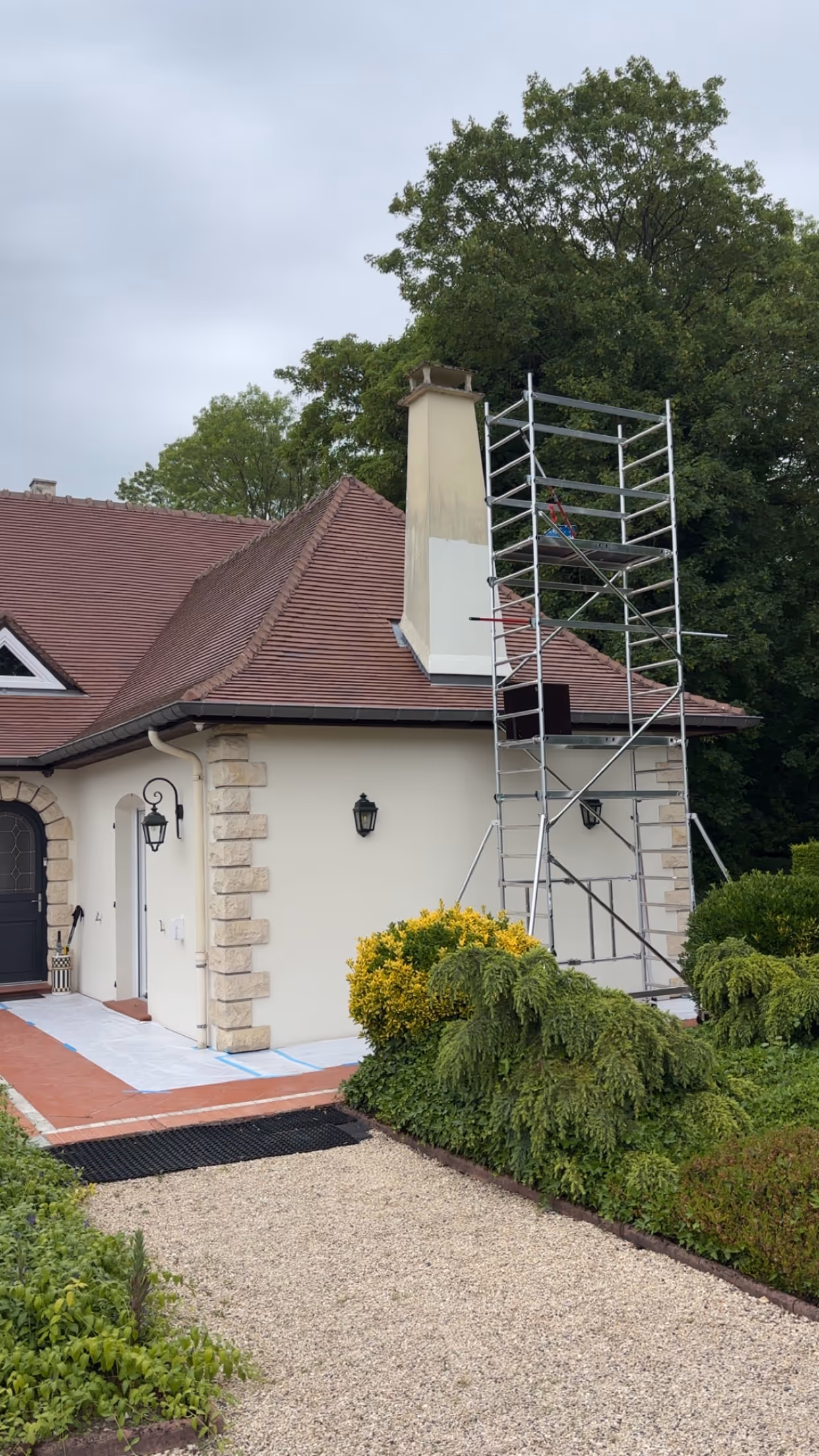 Maison avec un toit en tuiles rouges, une cheminée partiellement repeinte, un échafaudage, entourée d'arbustes verts et d'un chemin de gravier.