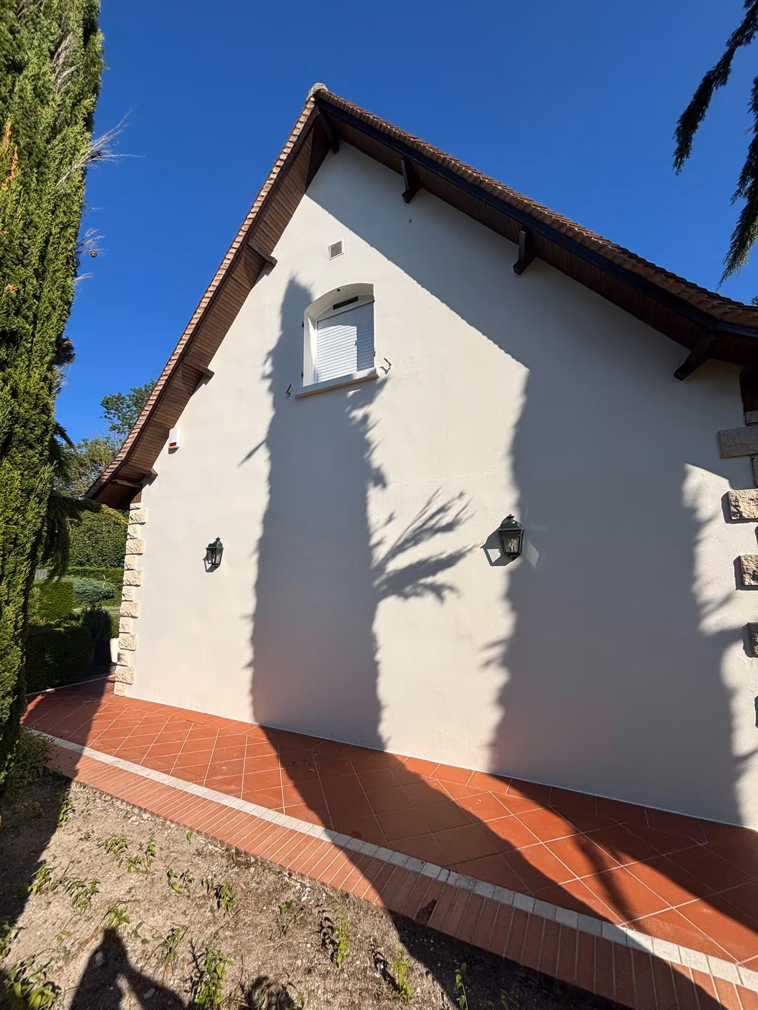 Façade blanche d'une maison sous un ciel bleu avec ombres d'arbres projetées, fenêtre fermée et lampes murales.