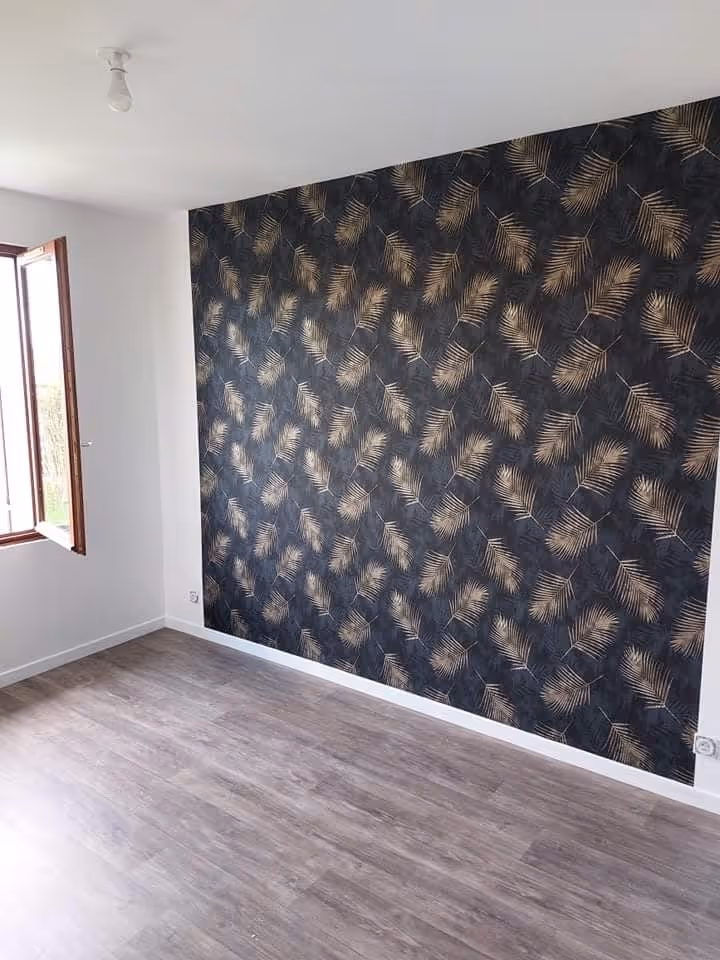 Chambre vide avec un mur d'accent décoré de papier peint noir à motif de feuilles dorées, un sol en bois clair, et une fenêtre ouverte.