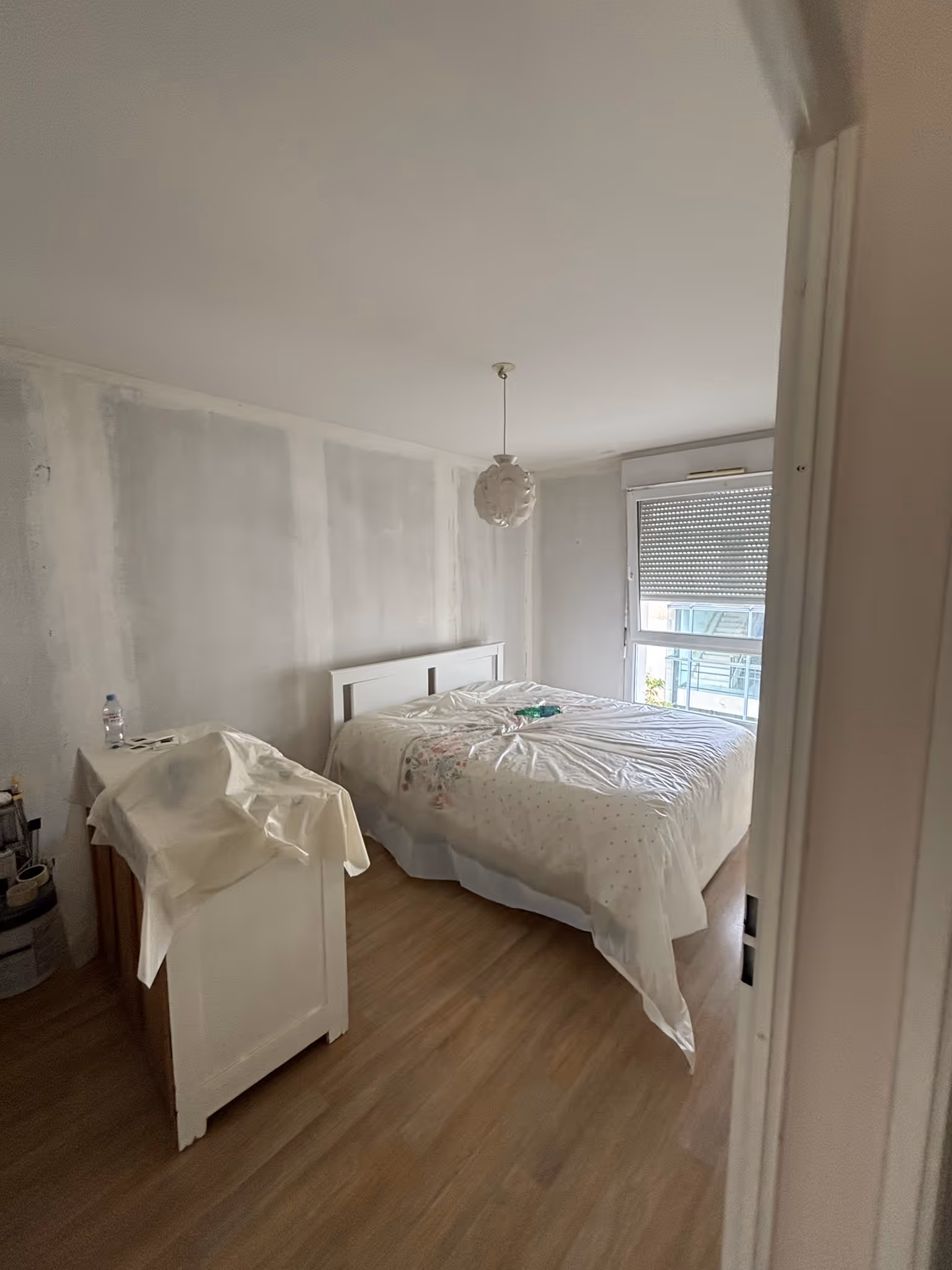 Chambre simple avec lit double, parquet clair, mur non peint, commode couverte d’un drap et fenêtre avec volet partiellement fermé.