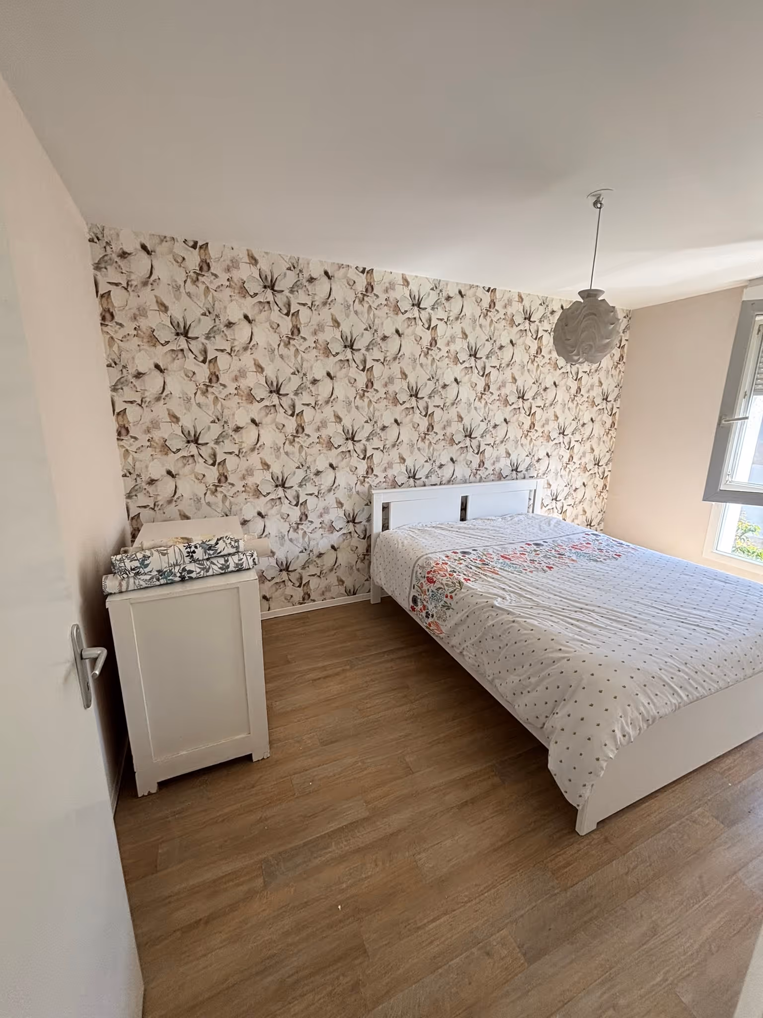 Chambre avec un lit double blanc, une commode blanche avec des tapis roulés dessus, parquet et mur à papier peint floral beige.