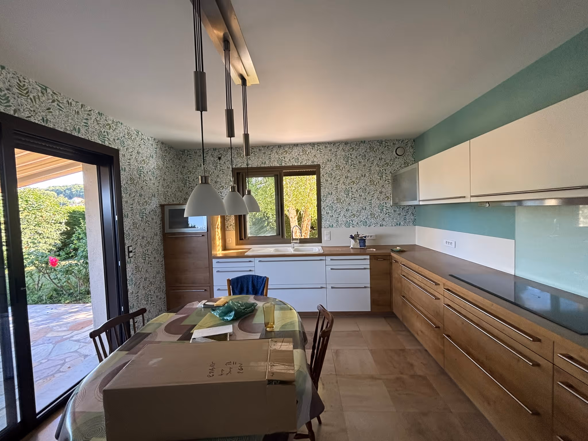Cuisine moderne avec des armoires en bois et blanches, une grande fenêtre, une table avec une nappe et un carton, et une porte vitrée donnant sur un jardin.
