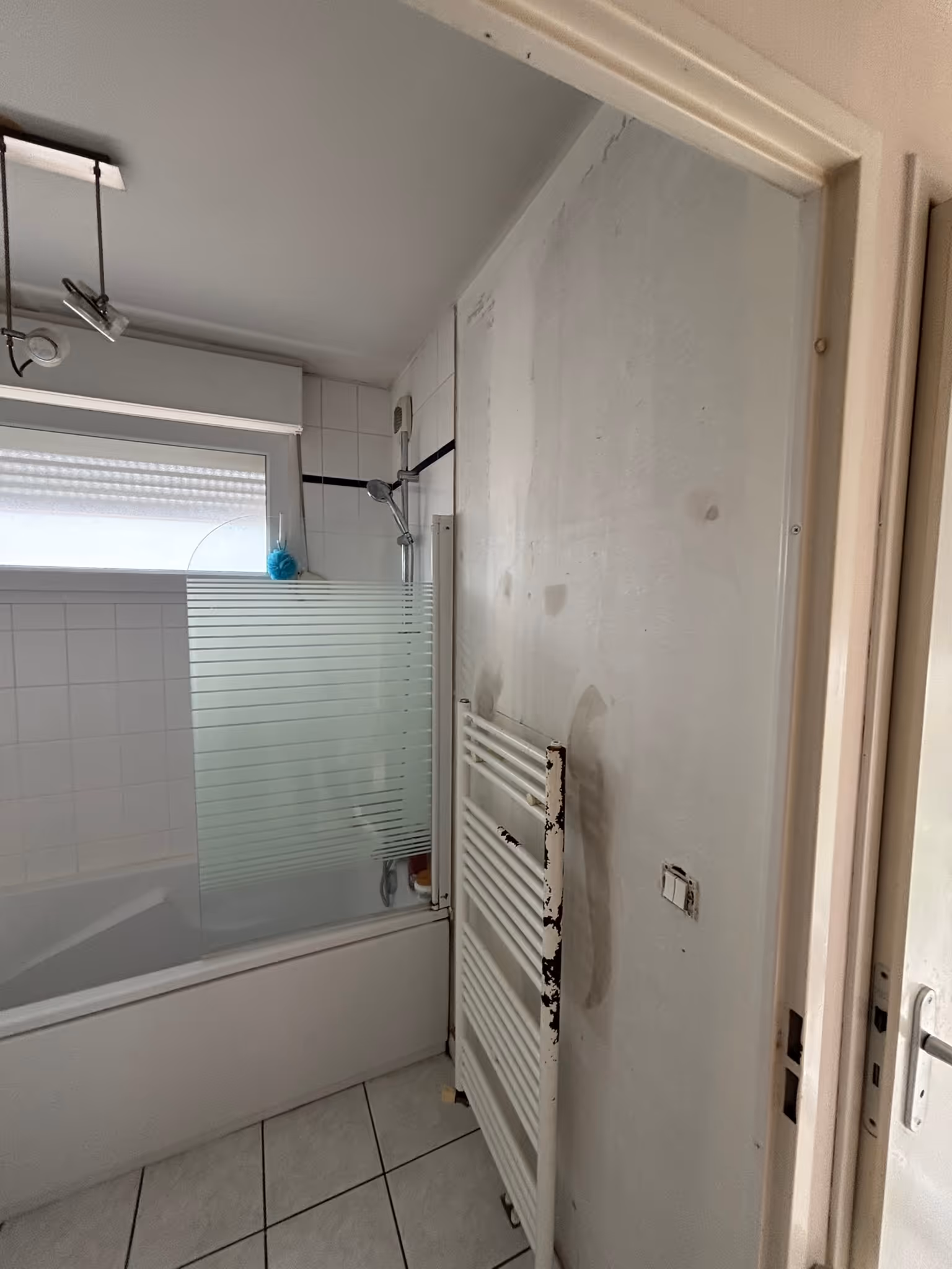 Salle de bains avec baignoire blanche, pare-douche partiellement givré, pomme de douche murale et radiateur sèche-serviettes avec peinture écaillée.