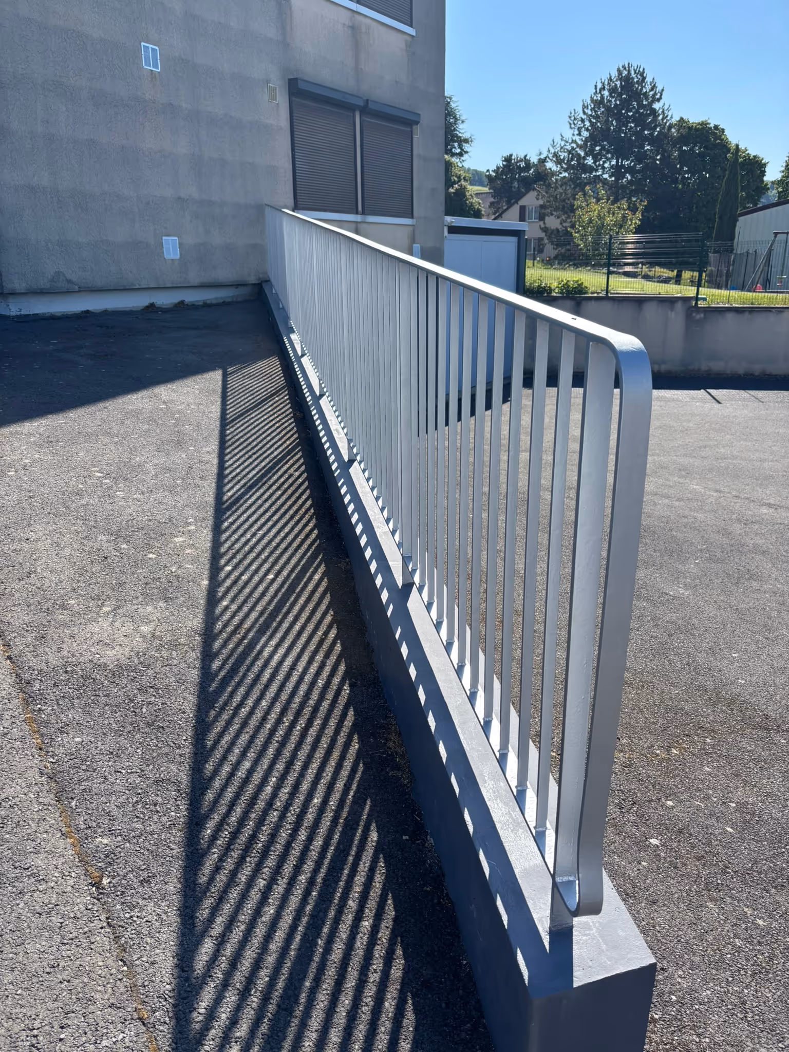 Rampe d'accès extérieure en métal avec ombres parallèles sur le sol asphalté, près d'un bâtiment gris.