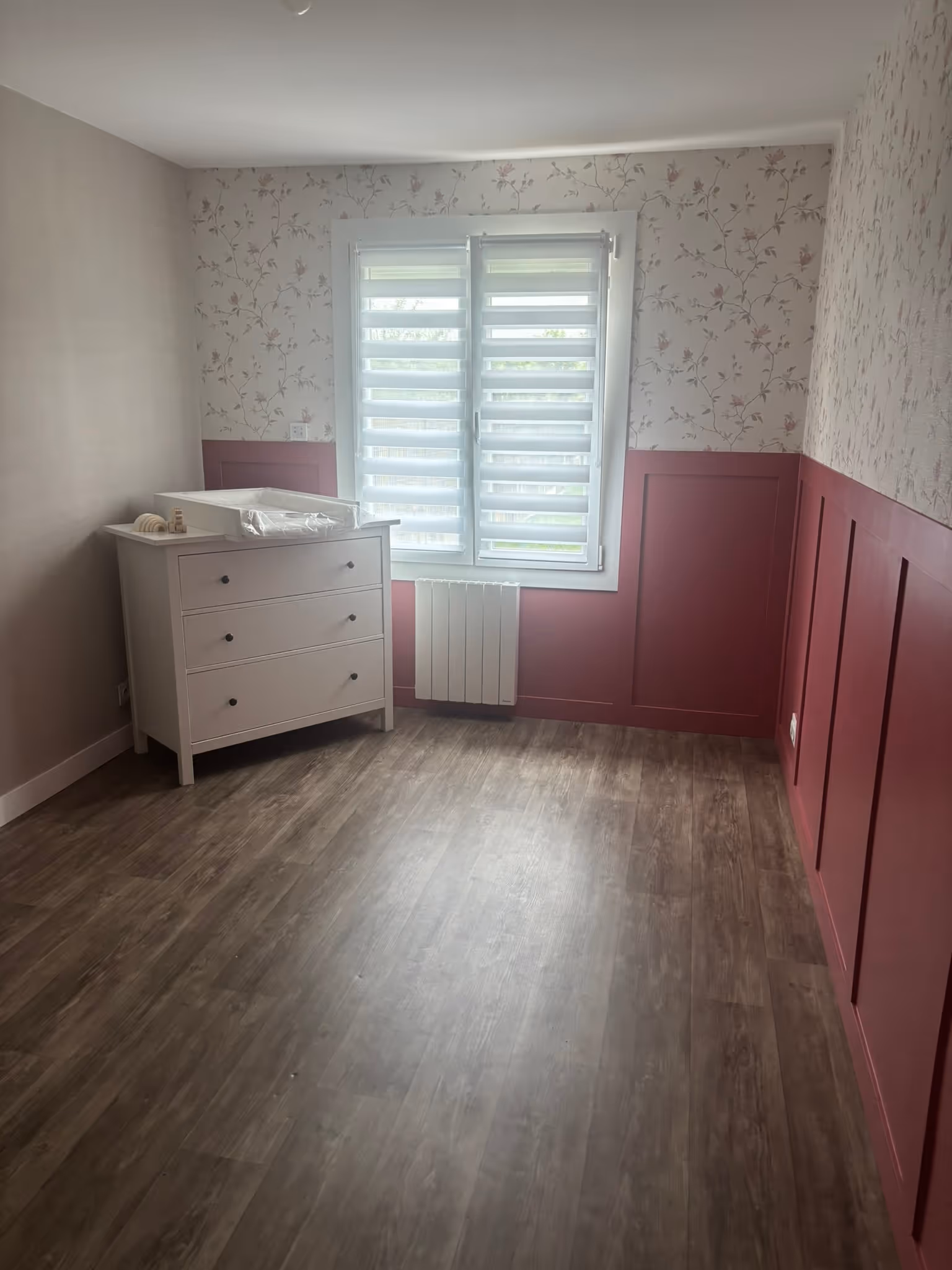 Chambre avec parquet foncé, murs moitié rose profond avec moulures et moitié papier peint floral, une commode blanche avec matelas à langer près d'une fenêtre à stores blancs.